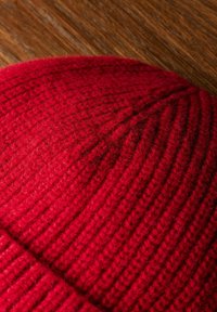 Bonnet en tricot rouge avec une texture côtelée et un matériau doux et duveteux. Forme incurvée avec un léger affaissement en haut, reposant sur un fond en bois.