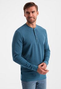Uomo sorridente, che indossa una maglietta henley blu a maniche lunghe con due bottoni, abbinata a jeans azzurri chiari, in piedi su uno sfondo semplice.