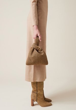 Borsa a tracolla marrone con manico annodato e dettaglio in anello dorato, abbinata a un vestito beige su misura e stivali al ginocchio coordinati.