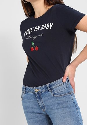 Kobieta w granatowym T-shircie z napisem "COME ON BABY Cherry me" i czerwonym haftem wiśni, w połączeniu z jasnoniebieskimi jeansami.