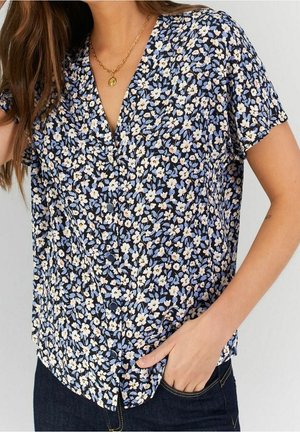 Kvinde iført kortærmet, knap-op bluse med små hvide og lyseblå blomsterprint, kombineret med mørke jeans og en guldfarvet vedhængs halskæde.