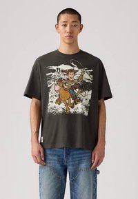 Νεαρός άνδρας φορεί σκούρο γκρι μπλουζάκι με κινουμένων σχεδίων καουμπόι που ιππεύει άλογο και κρατάει λούπες, σε συνδυασμό με μπλε τζιν.