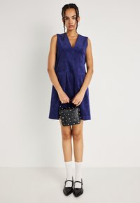 Fialové kordové šatové šaty bez rukávov s výstrihom do V, postrannými vreckami, kombinované s čiernou taškou s cvočkami, bielymi ponožkami s vlnkovaným okrajom a čiernymi topánkami.