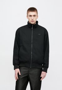 Μαύρο μπουφάν με φερμουάρ και ψηλό γιακά, με ριμπ μανσέτες· διαθέτει μαύρο λογότυπο και ριγέ διακοσμητικά στις μανσέτες· φορεμένο με γυαλιστερό σκούρο παντελόνι.