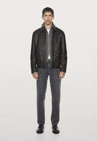 Giacca di pelle nera con colletto alto, due tasche frontali, abbinata a un maglione grigio, jeans grigi e scarpe nere su uno sfondo neutro.