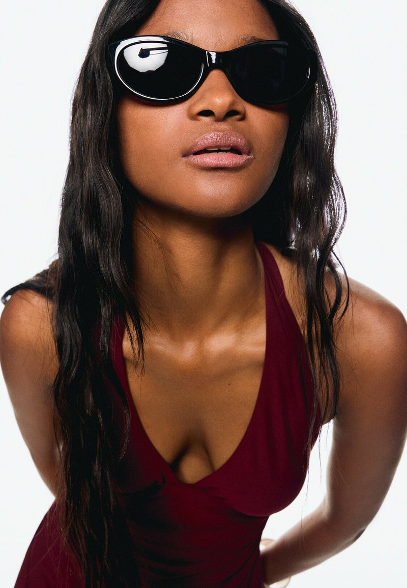 Black sunglasses with oversized frames, glossy finish. The model wears a deep burgundy dress with a V-neckline, highlighted by natural wavy hair.