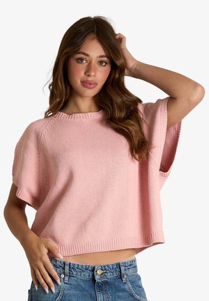 Giovane donna con lunghi capelli castani che indossa un top rosa a maniche corte e larghe lavorato a maglia e jeans blu, posando con una mano sulla testa e l'altra sul fianco.
