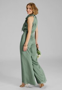 Groene wijde jumpsuit met een aansluitende bodice en een vloeiende striknek. Soepele stoftextuur, minimalistisch ontwerp, gecombineerd met gouden hakken.