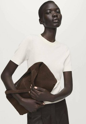 Brown suede tote bag held against a white knit short-sleeve shirt. The bag features a soft texture and a top handle. Simple design elements.