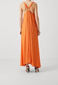 Robe maxi en tissu orange vif avec une coupe décontractée. Présente une découpe circulaire à l'arrière et des bretelles nouées. Ourlet bas avec mouvement.