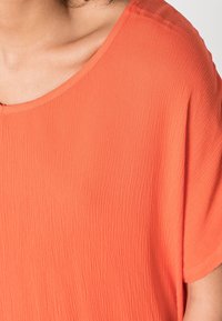 Korallenfarbene Bluse aus strukturiertem Stoff, mit weitem Halsausschnitt und lockerer Passform, kurzen Ärmeln und sanften Drapierungen.