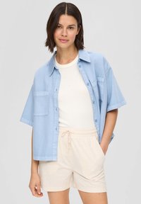 Lichtblauw shirt met korte mouwen, knopen aan de voorkant en borstzak, gedragen over een wit T-shirt en beige shorts met een elastische tailleband.