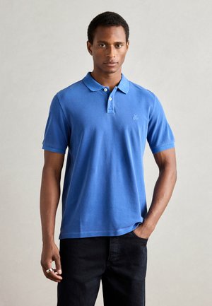 Polo shirt en coton bleu avec col et manches courtes, arborant un petit logo sur la poitrine, porté avec un jean noir. Texture lisse.
