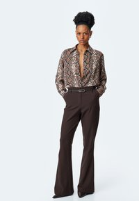 Blusa marrone con motivo serpente, con scollatura a V profonda, abbinata a pantaloni a vita alta marroni a campana e scarpe col tacco nere a punta.