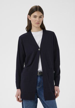 Femme portant un cardigan bleu marine à manches longues avec un seul bouton, un t-shirt blanc, un jean bleu et une ceinture noire sur un fond uni.
