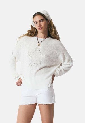 Un pull en tricot blanc avec un motif d'étoiles, porté avec des shorts en denim blanc ornés d'accents étoilés. Le modèle a les cheveux longs et porte un bandeau blanc.