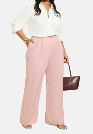 Femme portant une chemise blanche à boutons et un pantalon large taille haute rose clair, tenant un sac à main marron foncé dans une main.