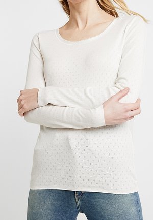Femme portant un haut blanc à manches longues avec de petites perforations en forme de losange et un jean bleu, les bras croisés à hauteur de la taille.