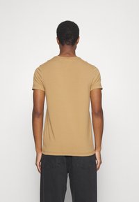 T-shirt ajusté à manches courtes en coton beige avec un col rond. Texture lisse, sans motifs. La vue arrière montre des détails subtils sur les coutures des épaules.