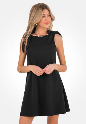 Kobieta w czarnej sukience bez rękawów z kokardkami na ramionach, stojąca ze splecionymi rękami, patrząca na bok na jednolitym tle.