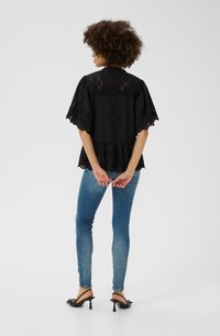Schwarzes Kurzarmshirt mit Lochstickerei und Rüsche am Saum, kombiniert mit eng anliegenden blauen Jeans. Das Modell trägt schwarze High Heels.