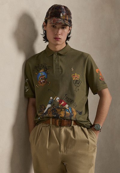 Πράσινο ελιάς πόλο με κεντημένους παίκτες πόλο, στέμμα και οικόσημο. Διαθέτει γιακά και κοντά μανίκια. Συνδυάζεται με μπεζ παντελόνια.