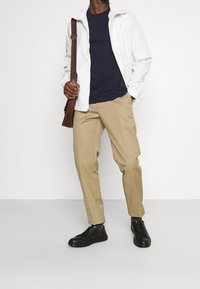 Valkoinen kevyt takki, tummansininen t-paita, beige housut ja mustat lenkkarit. Ruskea olkalaukku lisää kontrastia. Yksinkertainen, toiminnallinen muotoilu.