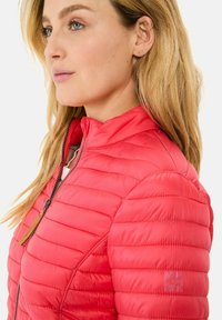 Rote wattierte Jacke mit Stehkragen, sichtbarem Reißverschluss und kleinem Logo am Ärmel. Das Material hat eine glatte, glänzende Textur.