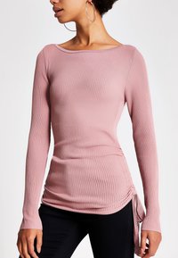 Langarm-Rippenoberteil in staubigem Rosa mit weitem Halsausschnitt, seitlichem Rüschen und Bändern. Das Material ist dehnbar und hat eine glatte Textur. Kombiniert mit schwarzen Hosen.