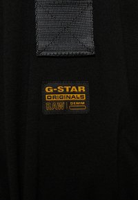 Czarny materiał z małą, wyszywaną prostokątną metką z napisem "G-STAR ORIGINALS RAW DENIM" w żółtej i czarnej nici.
