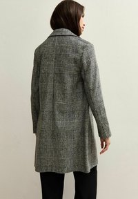 Abrigo de cuadros largo en gris y negro, con grandes patrones de cuadros, un cuello, mangas largas y una silueta alargada.