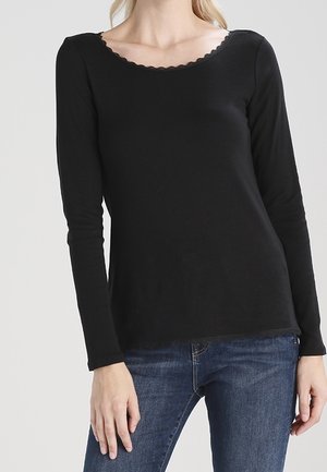 Mujer con camiseta negra de manga larga y escote con bordes ondulados, combinada con vaqueros azules, de pie frente a un fondo claro y liso.