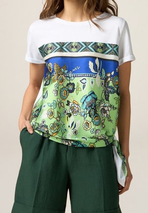 Kvinde iført en hvid T-shirt med et livligt blåt, grønt og blomsterprint foran, kombineret med mørkegrønne shorts, hånd i lommen.