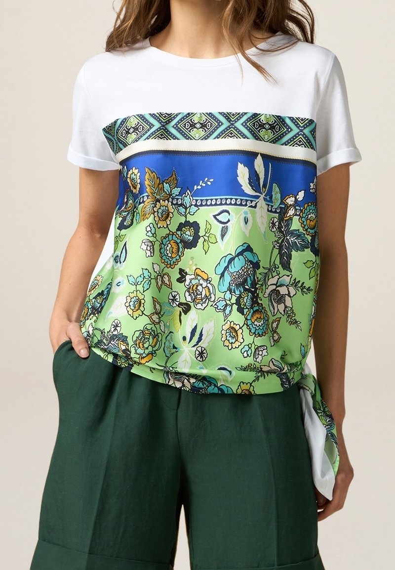 Femme portant un T-shirt blanc avec un panneau avant à motifs floraux bleu et vert vif, associé à un short vert foncé, main dans la poche.