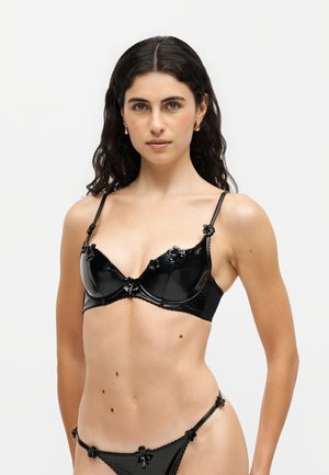 Femme aux cheveux bouclés foncés portant une lingerie noire brillante avec de petits détails floraux, debout devant un fond clair uni.