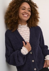 Mornarsko zložen cardigan z oversized rokavi, ki ima pet velikih okroglih gumbov in sproščen kroj, preden se nosi čez belo teksturirano bluzo.