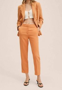 Orange skræddersyet jakkesæt med en enkeltknapjakke, slankebukser og en cropped hvid striktop med en nøglehulldetalje. Sorte sandaler.