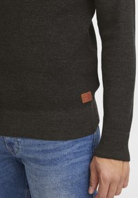 Mörkgrå stickad tröja med ribbade ärmslut och nederkant, med en liten brun läderetikett på nederkantens vänstra sida. Bärs över blåjeans.