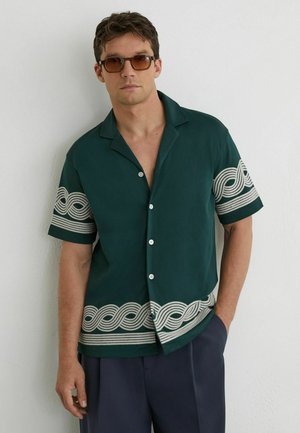 Bărbat purtând o cămașă verde închis cu mâneci scurte și nasturi, cu modele albe de valuri, pantaloni bleumarin și ochelari de soare maro, stând în fața unui perete alb.