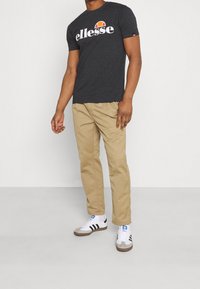 Camiseta de cuello redondo gris con un gran logo en blanco y naranja de "ellesse", combinada con pantalones beiges y zapatillas blancas con rayas negras.