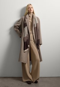 Beige wollen jas met een getailleerde pasvorm, gecombineerd met een bijpassende gebreide trui en wijde broek. Een patroon sjaal voegt textuur en contrast toe.