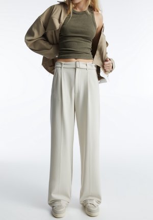 Model draagt beige brede broek met riem, bijpassend beige jasje, olijfgroen mouwloos geribd topje en beige sneakers tegen een witte achtergrond.