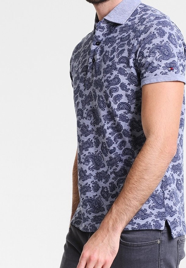 Homme portant un polo lavande à manches courtes avec un motif cachemire bleu foncé et un pantalon gris foncé sur un fond uni.