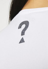 Camiseta blanca con un diseño de signo de interrogación y triángulo en purpurina metalizada plateada en la parte posterior, con una textura suave y agradable.