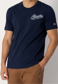 Muž v námořnicky modrém tričku Champion s bílým logem na hrudi a hnědých kalhotách s logem Champion na kapse, ruka v kapse.