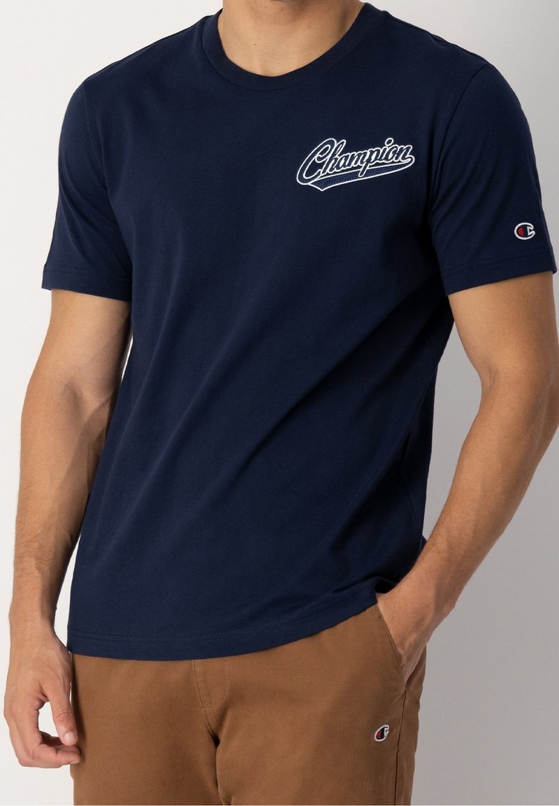 Muž v námořnicky modrém tričku Champion s bílým logem na hrudi a hnědých kalhotách s logem Champion na kapse, ruka v kapse.