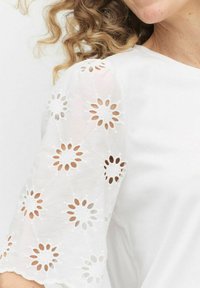 Vit blus med blommig broderad detalj på genomskinliga ärmar, med urklippta mönster och scallopade kanter för extra textur.