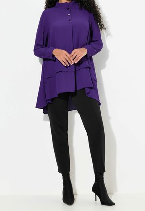 Kobieta ubrana w fioletową, piętrową, długą bluzkę z guzikami, czarne obcisłe spodnie i czarne botki na wysokim obcasie, stojąca na białym tle.