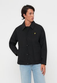 Veste noire avec un col, fermeture à boutons à l'avant et deux poches avant. Présente un petit logo jaune sur le côté gauche de la poitrine. Texture lisse.