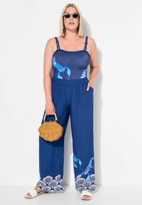 Námořnický modrý overal s potiskem humra a bílými akcenty, široké nohavice, nastavitelné ramínka a texturovaný pas. Zobrazený pletený košík.